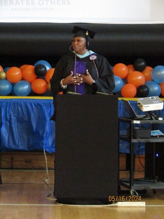 Adult in graduation robe speaking at a podium with balloons in background.