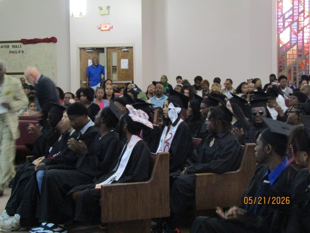 Audience, many in graduation attire, seated and clapping.