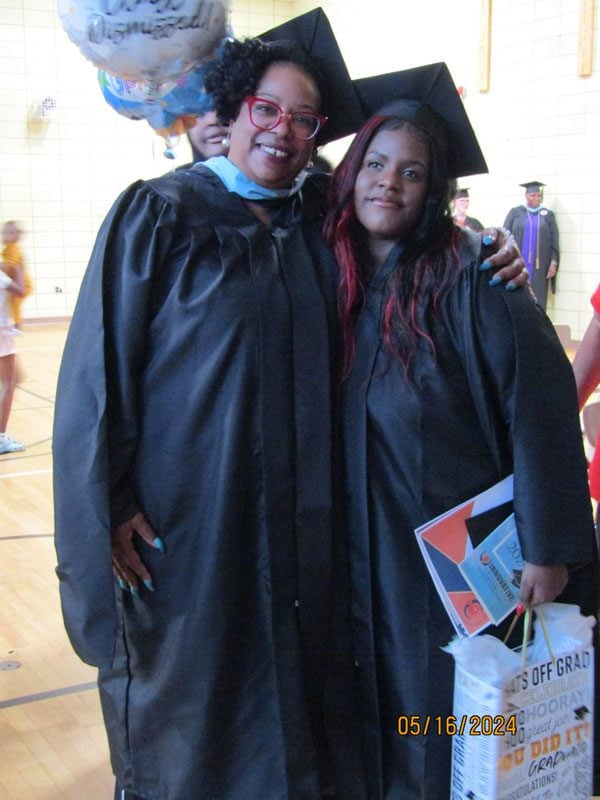 Two smiling adults in graduation attire pose, one holding a gift bag.