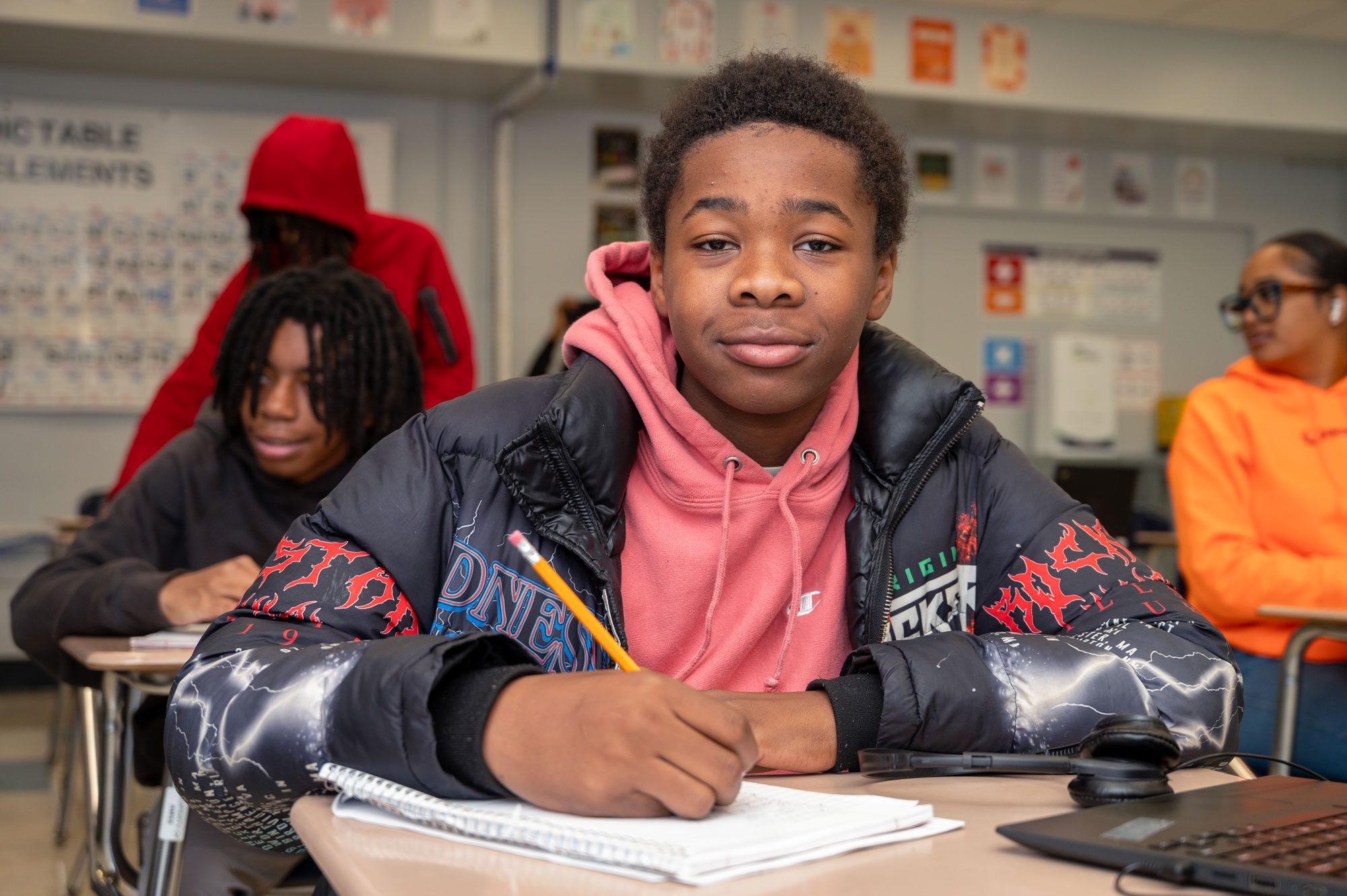 Student in a pink hoodie writing in a notebook at a classroom desk.